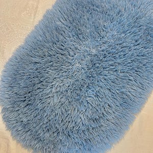 May include: A fluffy, oval-shaped, light blue rug. The rug has a dense, shaggy texture, with long, soft fibers creating a plush appearance. The color is a muted, pastel blue, giving it a cozy and inviting look.