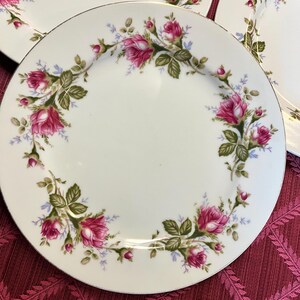 Puede incluir: Platos de cer&aacute;mica blanca con borde dorado y dise&ntilde;o floral de rosas. Los platos presentan un anillo de rosas rosas, hojas verdes y peque&ntilde;as flores azules alrededor del borde. Los platos est&aacute;n colocados sobre un mantel burdeos.