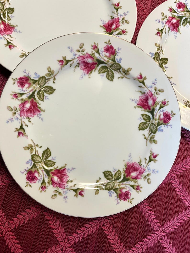 Puede incluir: Platos de cer&aacute;mica blanca con borde dorado y dise&ntilde;o floral de rosas. Los platos presentan un anillo de rosas rosas, hojas verdes y peque&ntilde;as flores azules alrededor del borde. Los platos est&aacute;n colocados sobre un mantel burdeos.
