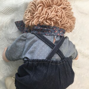 Puede incluir: Una mu&ntilde;eca hecha a mano con una peluca de lana marr&oacute;n claro, una camisa con estampado azul y blanco y un mono vaquero. La mu&ntilde;eca lleva zapatos blancos.