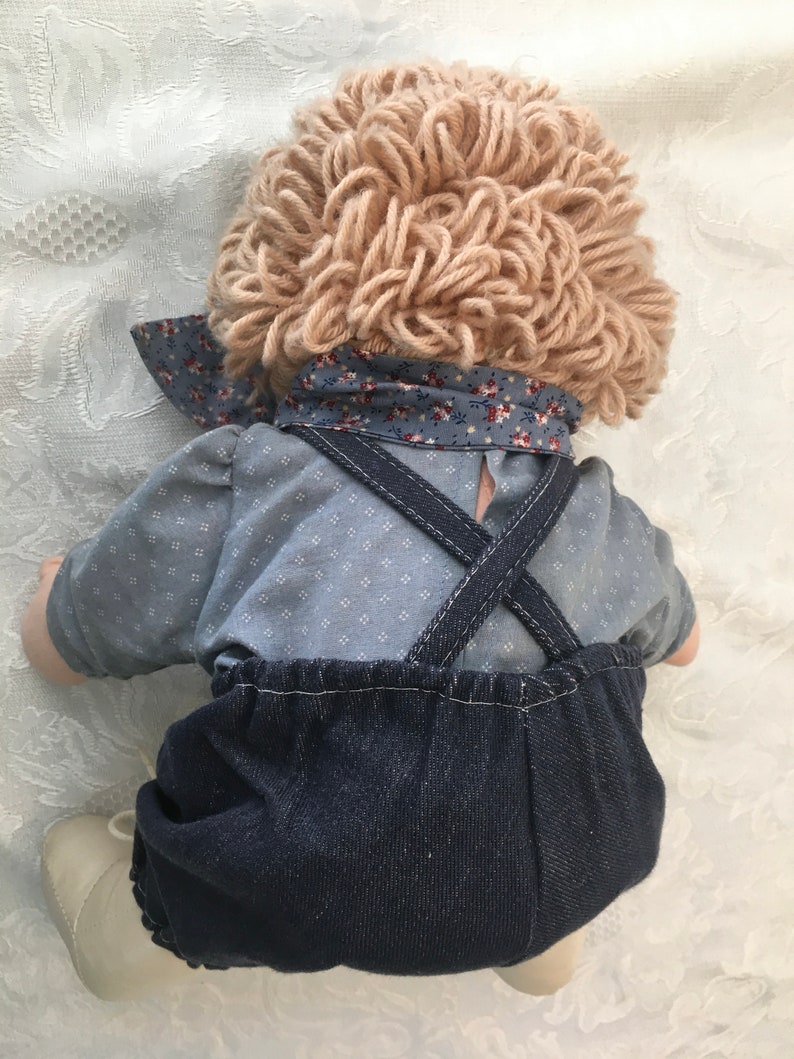 Puede incluir: Una mu&ntilde;eca hecha a mano con una peluca de lana marr&oacute;n claro, una camisa con estampado azul y blanco y un mono vaquero. La mu&ntilde;eca lleva zapatos blancos.