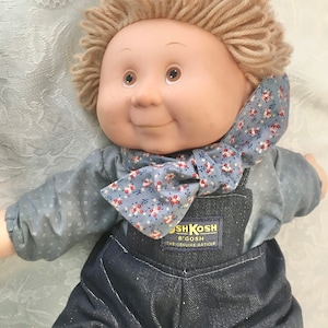 Puede incluir: Una mu&ntilde;eca de peluche Cabbage Patch Kids con pelo de lana rubio, que lleva una camisa de patr&oacute;n azul y blanco y un mono azul. La mu&ntilde;eca tiene una pajarita floral azul y blanca y una etiqueta de OshKosh B'Gosh en el mono.