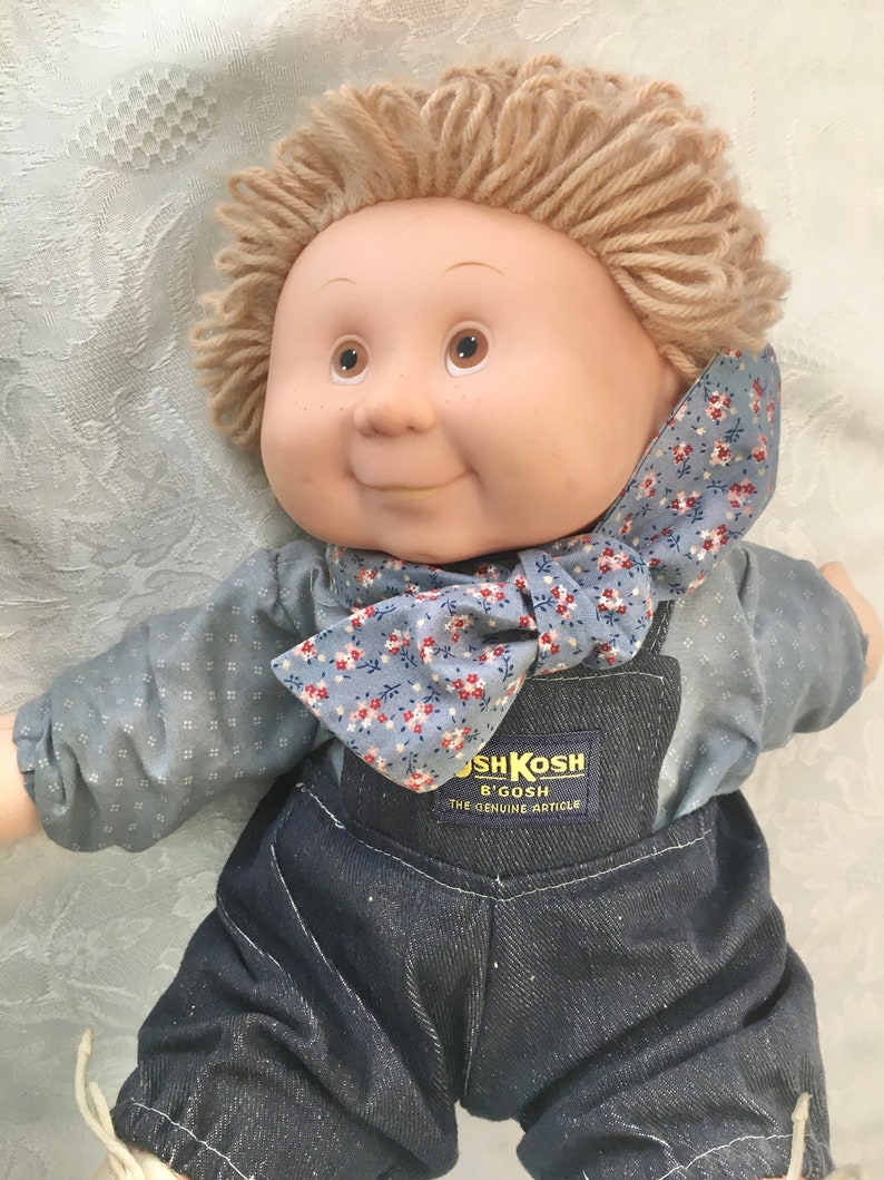Puede incluir: Una mu&ntilde;eca de peluche Cabbage Patch Kids con pelo de lana rubio, que lleva una camisa de patr&oacute;n azul y blanco y un mono azul. La mu&ntilde;eca tiene una pajarita floral azul y blanca y una etiqueta de OshKosh B'Gosh en el mono.