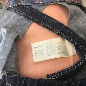 Puede incluir: Una mu&ntilde;eca de tela azul y blanca con un babero de mezclilla y una etiqueta que dice "Blue Box" y "Contenido: Todos los materiales nuevos Relleno de fibra de poli&eacute;ster OH-1073 PA-1073 MA-T-533-M Hecho en Hong Kong".