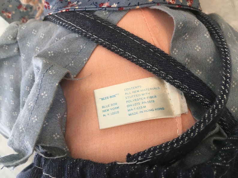 Puede incluir: Una mu&ntilde;eca de tela azul y blanca con un babero de mezclilla y una etiqueta que dice "Blue Box" y "Contenido: Todos los materiales nuevos Relleno de fibra de poli&eacute;ster OH-1073 PA-1073 MA-T-533-M Hecho en Hong Kong".