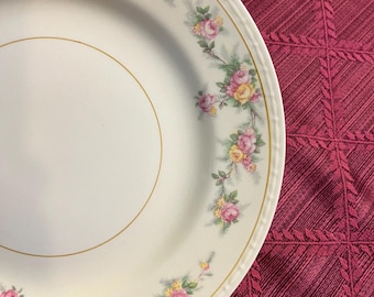 May include: A white ceramic plate with a gold rim and a floral pattern of pink and yellow roses. The plate has a scalloped edge and is set against a burgundy background.