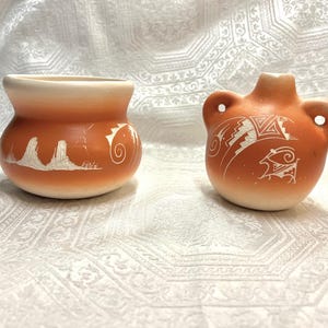 May include: Two terracotta-colored ceramic vessels with white etched designs. The left piece is a wide-mouthed pot featuring a landscape scene. The right piece is a rounded canteen-style vessel with two side handles and geometric animal motifs.