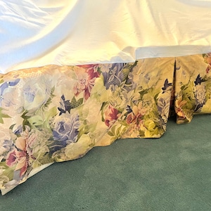 May include: A floral bed skirt with a cream background and a pattern of pink, purple, and blue flowers. The bed skirt is pleated and has a scalloped edge. The bed skirt is made of a soft, silky fabric.