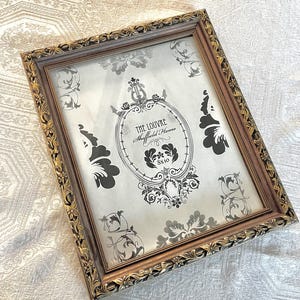 May include: Rectangular picture frame with ornate gold trim and a brown inner border. The frame holds a print with a black and white floral design and the text "THE LOUVRE Sheffield Home 8x10".
