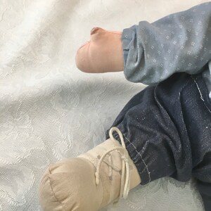 Puede incluir: Primer plano de un pie de mu&ntilde;eca con un zapato marr&oacute;n claro con cordones blancos. La mu&ntilde;eca lleva pantalones vaqueros azules y una camisa azul claro con un patr&oacute;n blanco.