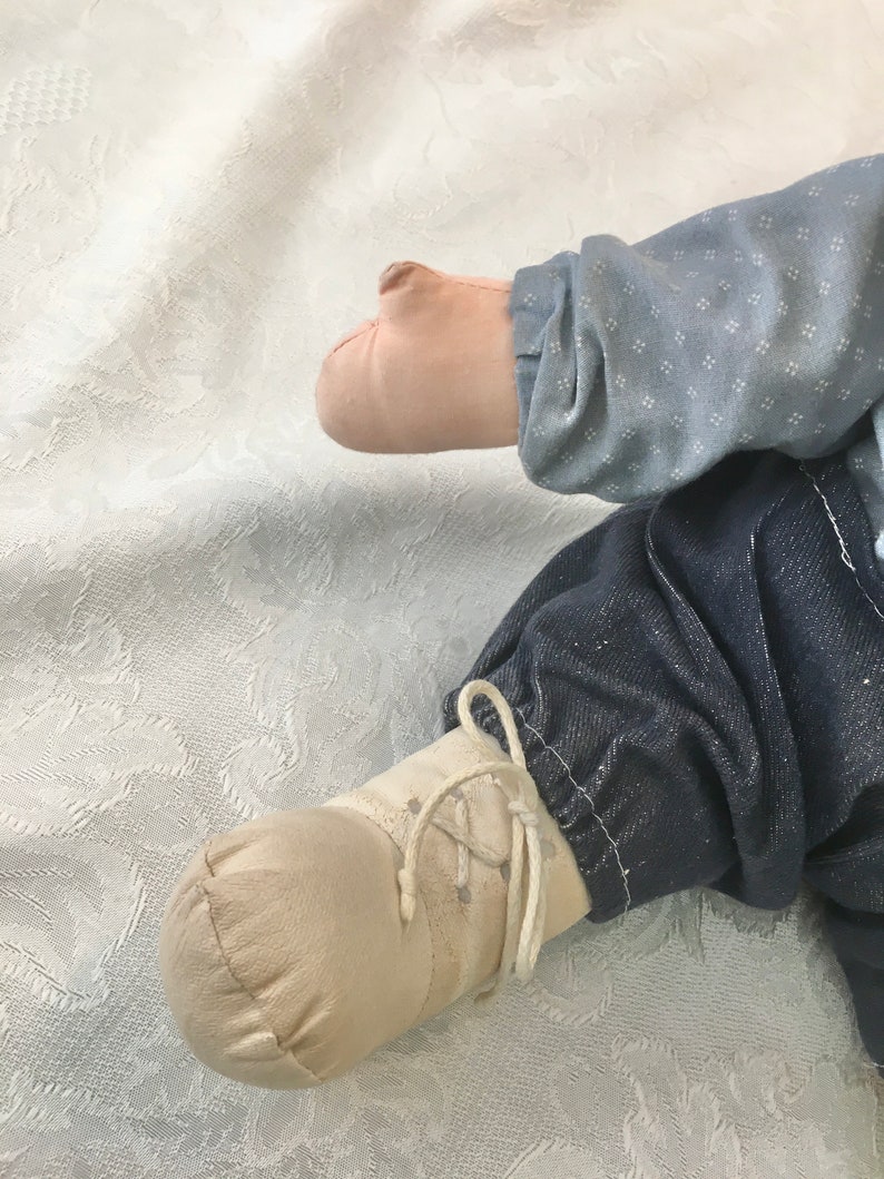 Puede incluir: Primer plano de un pie de mu&ntilde;eca con un zapato marr&oacute;n claro con cordones blancos. La mu&ntilde;eca lleva pantalones vaqueros azules y una camisa azul claro con un patr&oacute;n blanco.