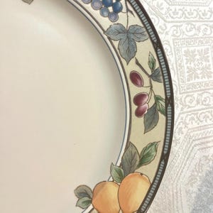 May include: A white ceramic plate with a decorative border featuring illustrations of fruit, including grapes and oranges. The border has a light beige background with a blue and brown trim. The plate is partially visible.