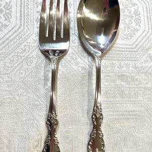 May include: A silver serving fork and spoon set. The fork has four tines, and the spoon has a large, oval bowl. Both utensils have ornate floral detailing on the handles, and are made of a shiny, reflective metal.