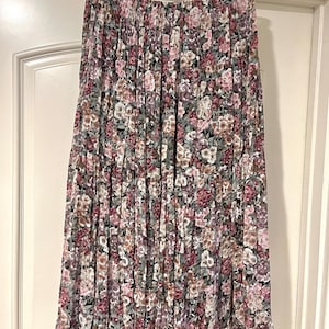 May include: A floral print skirt with a tiered design. The skirt features a mix of pink, purple, and white flowers on a black background. The skirt is hanging on a white hanger against a white door.