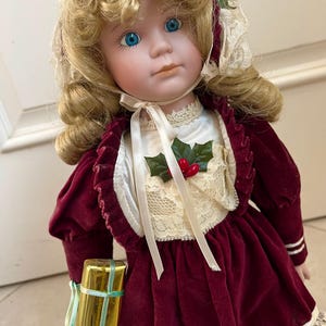 May include: A porcelain doll with curly blonde hair and blue eyes, wearing a deep red velvet dress with a cream-colored lace bodice, a matching bonnet with lace trim, and a holly sprig accent. The doll holds a small gold gift box.