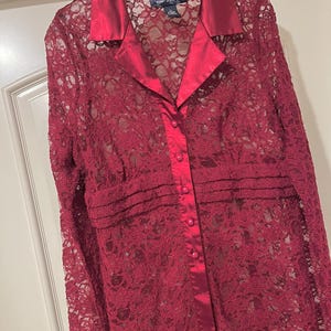 May include: A burgundy lace shirt with a satin collar and button-up front. The long-sleeved shirt is hanging on a white hanger. The shirt has a floral lace pattern.