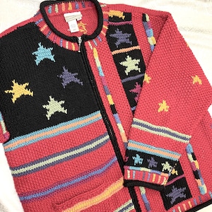May include: A red zip-up sweater with a black front panel adorned with yellow, blue, and green star designs. The sweater features colorful striped accents and a black collar and cuffs. The sweater is made of a knitted material.