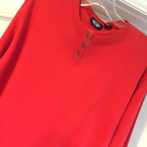 May include: A long-sleeved, red, button-up shirt with a round neckline. The shirt has a small logo patch on the left side of the chest that says "Wings of Freedom".