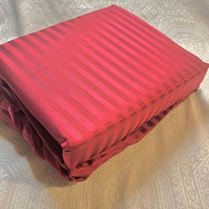 May include: Folded red bed sheet set with a striped pattern. The sheets are a deep red color and appear to be made of a smooth, possibly satin, material. The stripes run vertically across the fabric.