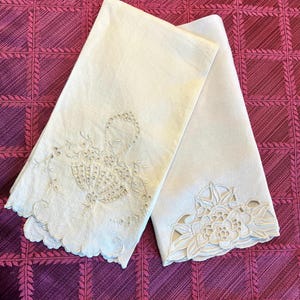 May include: Two white linen tea towels with delicate embroidered floral designs. One towel features a basket of flowers, while the other has a single large flower.
