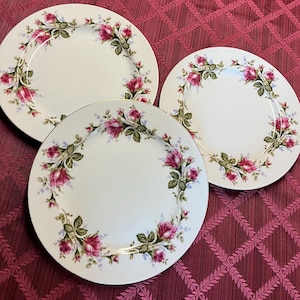 Puede incluir: Tres platos de cer&aacute;mica blanca con un dise&ntilde;o floral de rosas. Los platos tienen un borde dorado y un borde de rosas rosas, hojas verdes y peque&ntilde;as flores azules. Los platos est&aacute;n sobre un mantel burdeos.