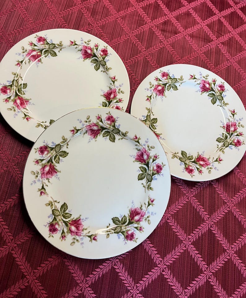 Puede incluir: Tres platos de cer&aacute;mica blanca con un dise&ntilde;o floral de rosas. Los platos tienen un borde dorado y un borde de rosas rosas, hojas verdes y peque&ntilde;as flores azules. Los platos est&aacute;n sobre un mantel burdeos.