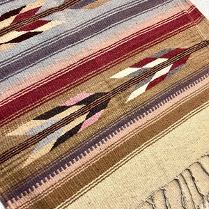 May include: A woven throw blanket with a southwestern pattern featuring brown, beige, pink, red, and gray stripes with arrow designs. The blanket has a fringe border.