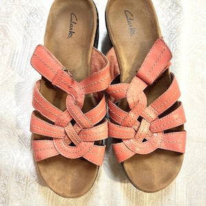 May include: A pair of coral-colored leather sandals with a woven strap design across the top. The sandals feature a tan contoured footbed with the Clarks brand name printed in black on the heel area and an adjustable hook-and-loop closure strap.