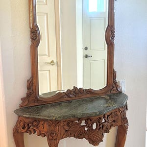 May include: A wooden console table with a dark green marble top and intricate floral carvings on the legs and apron, paired with a matching tall arched wall mirror featuring elaborate wood-carved scrollwork and a central decorative motif.