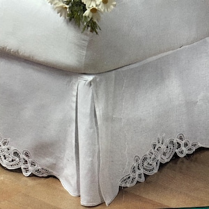May include: White bed skirt with a delicate lace trim along the bottom edge. The skirt has a pleated design and is draped over a bed frame. A small bouquet of white flowers rests on the bed.