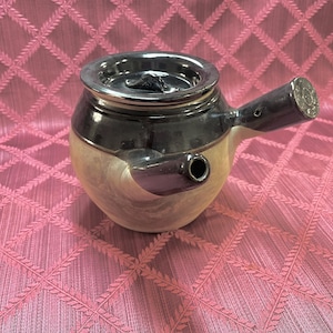May include: A ceramic teapot with a silver lid and spout. The teapot has a dark brown top and a light beige body. The spout is cylindrical and angled. The teapot is sitting on a pink patterned tablecloth.