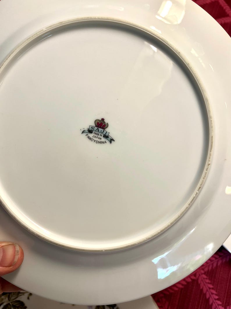 Puede incluir: Plato de porcelana blanca con un borde ligeramente elevado. La parte inferior presenta un emblema decorativo con el texto "SAJI MADE IN JAPAN FANCYCHINA" en negro y rojo. El plato est&aacute; limpio y parece sin usar.