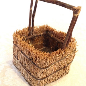 May include: A small, square-shaped basket made of woven straw with a wooden handle. The basket is brown and has a natural, rustic look.