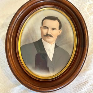 May include: An oval portrait of a man with a mustache, wearing a dark suit and white collared shirt. The portrait is encased in a polished dark brown wooden frame with a thin gold-toned inner border, featuring a soft, faded background.