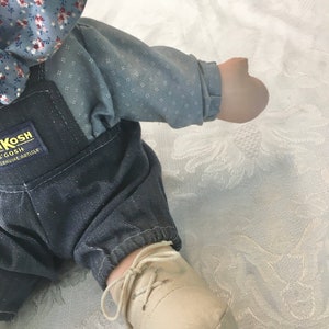 Puede incluir: Una mu&ntilde;eca de tela azul y blanca con un mono vaquero y un zapato blanco con un cord&oacute;n marr&oacute;n. La mu&ntilde;eca tiene un lazo de tela azul y blanca en la cabeza. La etiqueta de la mu&ntilde;eca dice "Kosh  Genuine Article".