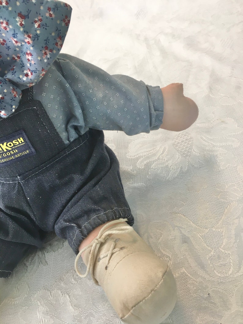 Puede incluir: Una mu&ntilde;eca de tela azul y blanca con un mono vaquero y un zapato blanco con un cord&oacute;n marr&oacute;n. La mu&ntilde;eca tiene un lazo de tela azul y blanca en la cabeza. La etiqueta de la mu&ntilde;eca dice "Kosh  Genuine Article".