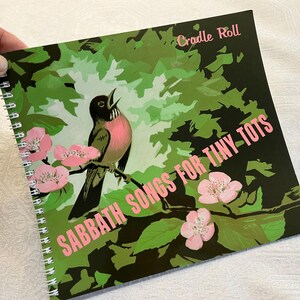 May include: A spiral bound book with a green, black, and pink cover. The cover features a bird singing in a tree with pink flowers. The title of the book is "Sabbath Songs for Tiny-Tots" and "Cradle Roll" is printed in the top right corner.