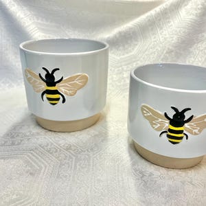 May include: Two white ceramic planters with a bee design. The bee is black and yellow with brown wings. The planters have a beige base.