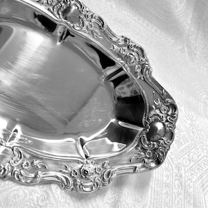 May include: A silver-colored, ornate serving tray with a highly reflective surface. The tray has a detailed, floral-inspired border design. The background is a white, patterned fabric.