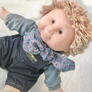 Puede incluir: Una mu&ntilde;eca Cabbage Patch Kids con pelo de lana rubio que lleva una camisa de estampado floral azul y blanco y un mono. El mono tiene el texto "BIGOSH" y "THE GENUINE ARTICLE" impreso.