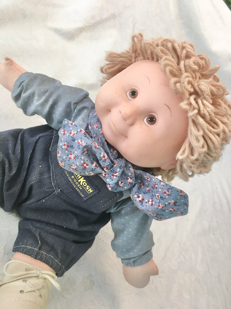 Puede incluir: Una mu&ntilde;eca Cabbage Patch Kids con pelo de lana rubio que lleva una camisa de estampado floral azul y blanco y un mono. El mono tiene el texto "BIGOSH" y "THE GENUINE ARTICLE" impreso.
