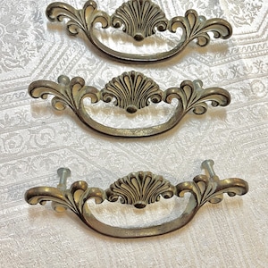 May include: Three antique brass drawer pulls with a shell and scroll design. The pulls have a curved handle and are mounted with screws. The hardware has a vintage, aged appearance, suggesting they are repurposed or vintage.