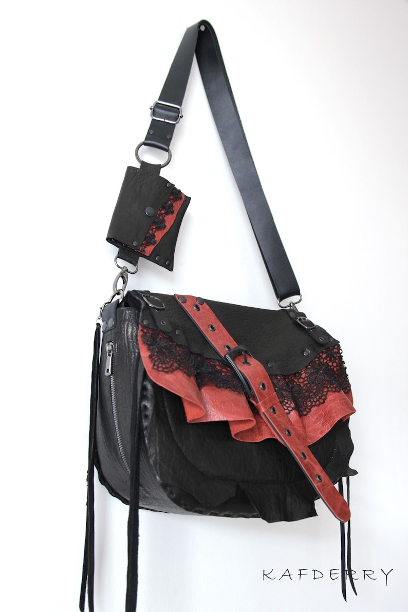 K&ouml;nnte beinhalten: Schwarze Leder-Umh&auml;ngetasche mit roten Akzenten und Spitzenbesatz. Mit einer passenden kleinen Tasche und einem verstellbaren Riemen. Die Tasche hat einen Schnallenverschluss und dekorative Quasten.