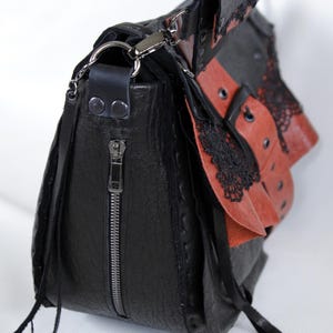 K&ouml;nnte beinhalten: Schwarze Leder-Umh&auml;ngetasche mit roten Akzenten und schwarzer Spitzenborte. Die Tasche hat einen silbernen Rei&szlig;verschluss, einen langen Riemen und Quasten. Das Design umfasst mehrere Schichten und Texturen, mit dem Markennamen "KAFDERY" sichtbar.