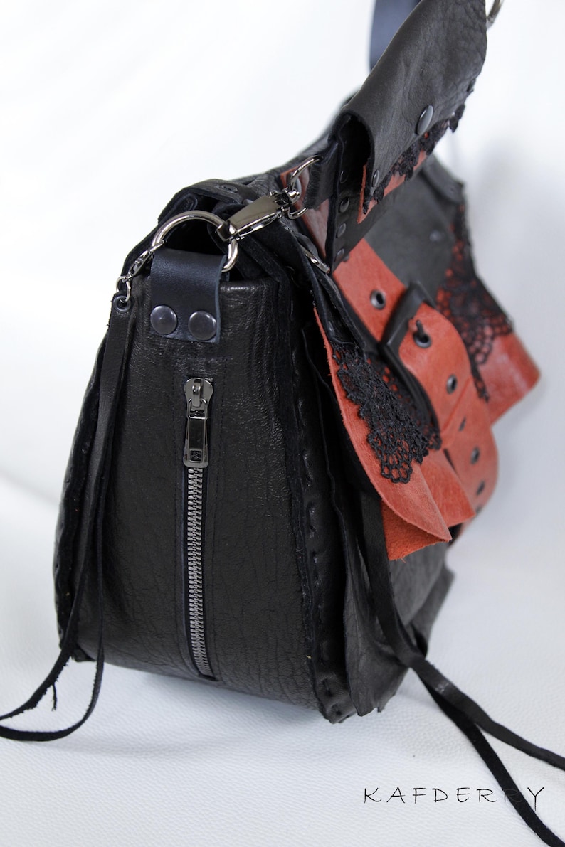 K&ouml;nnte beinhalten: Schwarze Leder-Umh&auml;ngetasche mit roten Akzenten und schwarzer Spitzenborte. Die Tasche hat einen silbernen Rei&szlig;verschluss, einen langen Riemen und Quasten. Das Design umfasst mehrere Schichten und Texturen, mit dem Markennamen "KAFDERY" sichtbar.