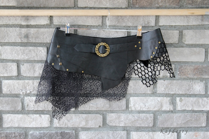 May include: A black asymmetrical skirt featuring a wide faux leather waistband with a brass-toned circular buckle, metallic studs, and a layered black mesh overlay with a textured, web-like pattern hanging from the hemline.