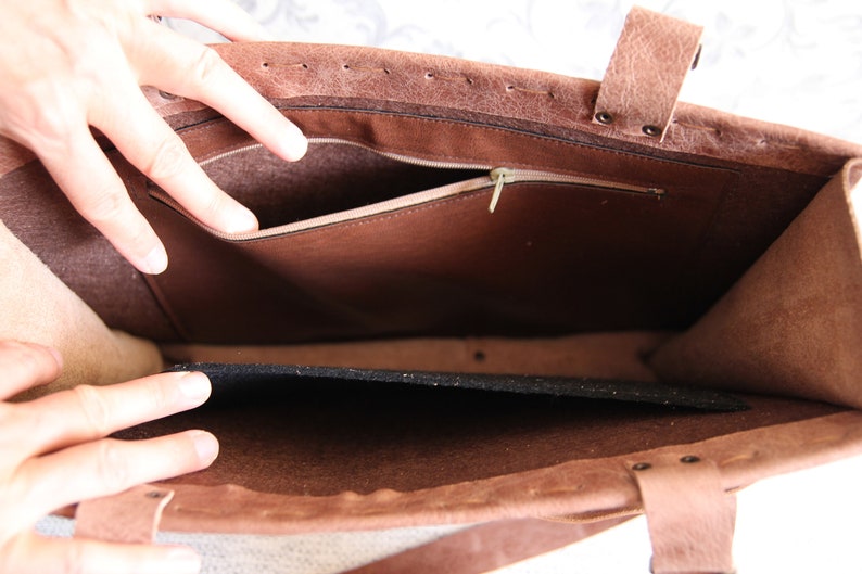 May include: A brown leather tote bag with a textured exterior, featuring a spacious interior compartment, a zippered pocket along the inner wall, and a removable black felt insert at the base. The bag is constructed with visible stitching.