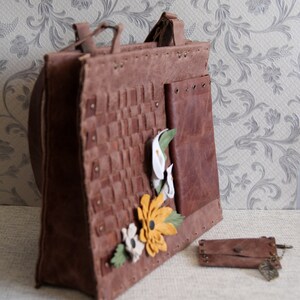 May include: A brown leather tote bag featuring a woven front panel, a smooth side pocket with metal grommets, and decorative leather flower appliqu&eacute;s in white and yellow. A matching small leather pouch with a leaf charm sits nearby. Text reads Kafderry.
