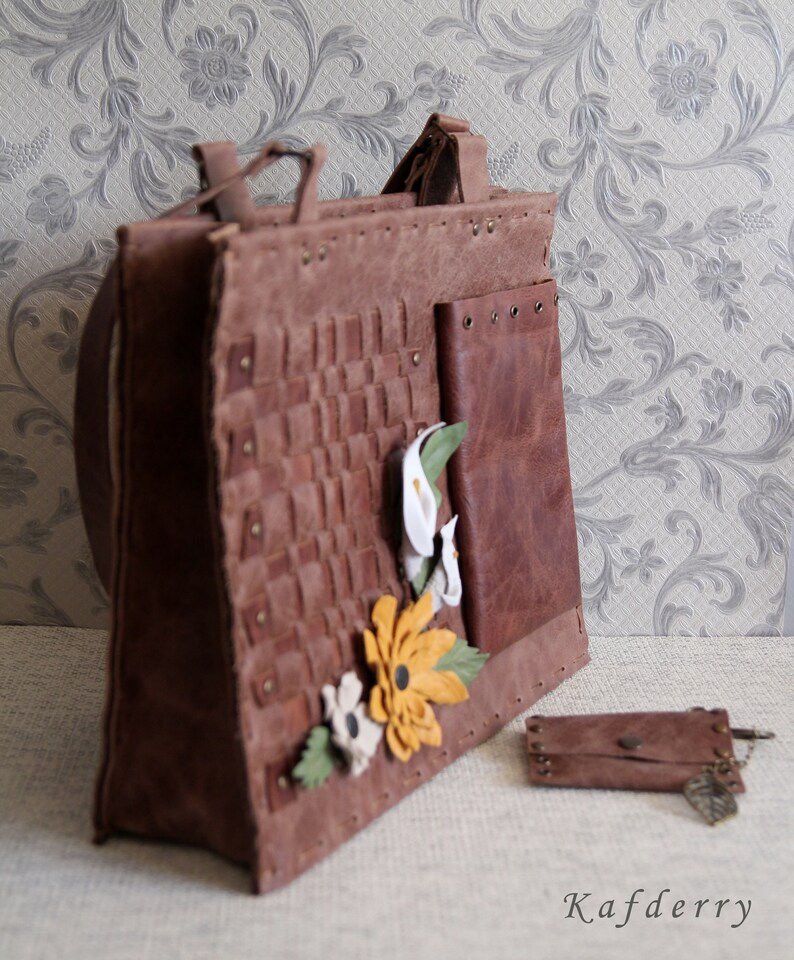 May include: A brown leather tote bag featuring a woven front panel, a smooth side pocket with metal grommets, and decorative leather flower appliqu&eacute;s in white and yellow. A matching small leather pouch with a leaf charm sits nearby. Text reads Kafderry.
