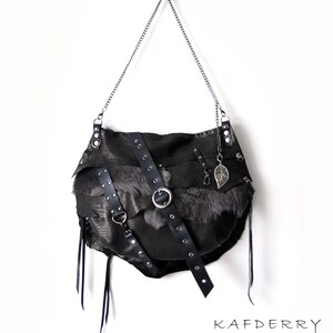 Puede incluir: Bolso de hombro de cuero negro con correa de cadena, con un panel de piel y correas decorativas con herrajes plateados. Un dije de hoja plateada cuelga de la cadena. El bolso tiene un dise&ntilde;o asim&eacute;trico &uacute;nico. La marca "KAFDERRY" est&aacute; impresa.